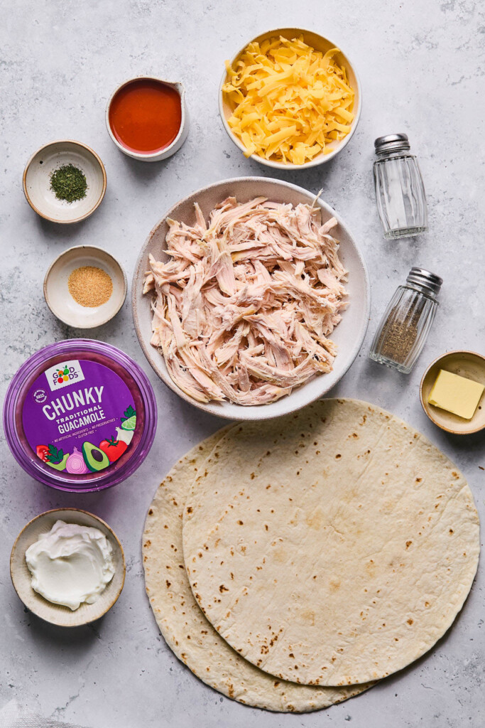 Grey concrete counter with a bowl of shredded cheddar cheese, a salt and pepper shaker, a small dish with some butter in it, two large tortillas, a bowl of greek yogurt, a container of good foods chunky guacamole, a small dish of garlic powder, a small dish of dried dill, and a cup of buffalo sauce.