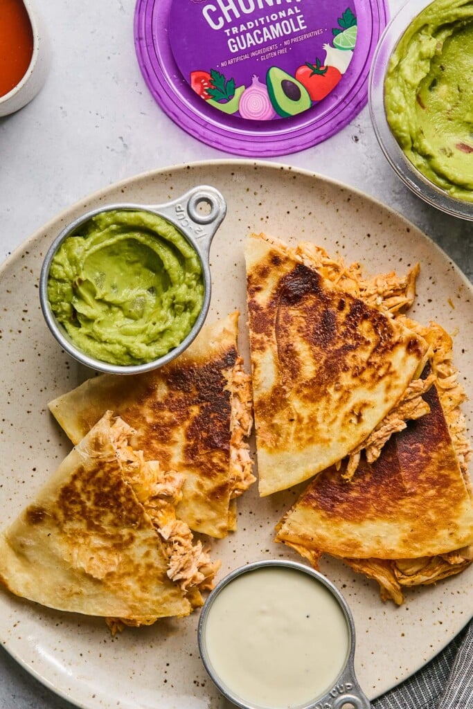 Buffalo chicken quesadillas on a plate with a small dish of guacamole and a small dish of blue cheese dressing. Next to the plate is a large container of guacamole and a small dish of buffalo sauce.