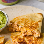 A plate with pieces of buffalo chicken quesadilla on it and a small dish of guacamole. Behind the plate is a large container of guacamole.