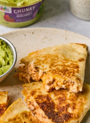A plate with pieces of buffalo chicken quesadilla on it and a small dish of guacamole. Behind the plate is a large container of guacamole.