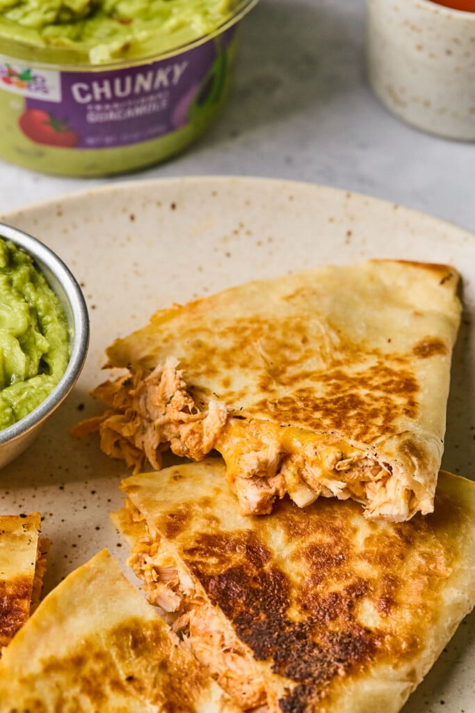 A plate with pieces of buffalo chicken quesadilla on it and a small dish of guacamole. Behind the plate is a large container of guacamole.