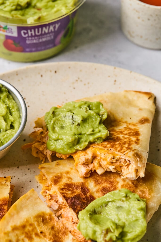 A close up of a plate with pieces of buffalo chicken quesadillas on it topped with guacamole. There is a small dish of guacamole on the plate as well. Behind the plate is a large container of guacamole.