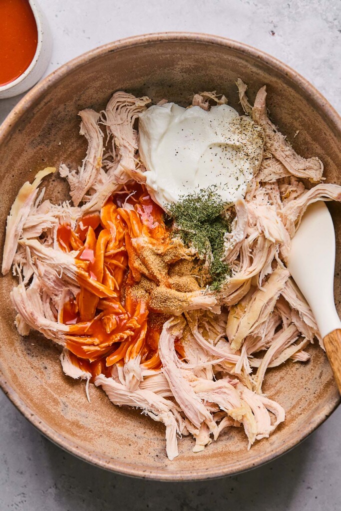 A bowl of shredded chicken, hot sauce, greek yogurt, and spices with a spatula in it. Next to the bowl is a small cup of more hot sauce.