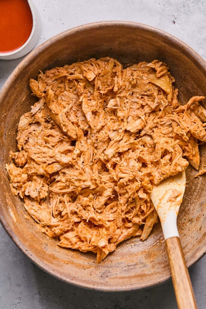 A bowl with shredded buffalo chicken in it with a spatula in it. Next to the bowl is a small cup of hot sauce.
