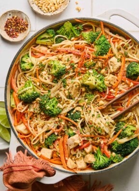 Peanut butter noodles with chicken and veggies in a large white skillet. It is garnished with cilantro and chopped peanuts and also in the pan is a pair of chopsticks. The handle of the pan is wrapped in a red towel and around the pan is a plate of lime wedges, a bowl of red pepper flakes, and a bowl of chopped peanuts.