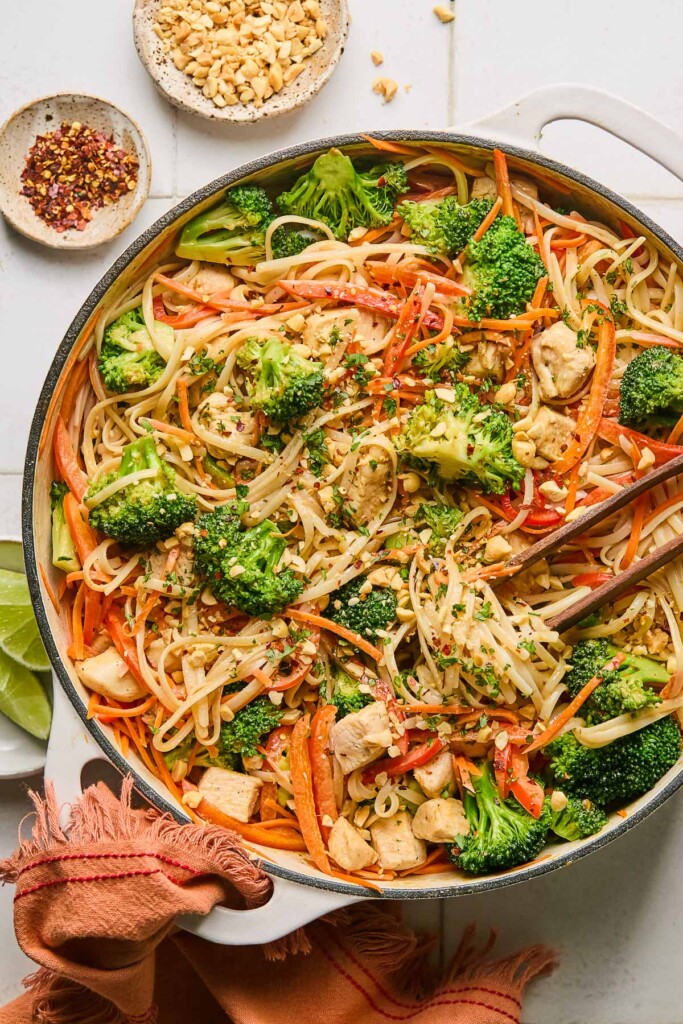 Peanut butter noodles with chicken and veggies in a large white skillet. It is garnished with cilantro and chopped peanuts and also in the pan is a pair of chopsticks. The handle of the pan is wrapped in a red towel and around the pan is a plate of lime wedges, a bowl of red pepper flakes, and a bowl of chopped peanuts.