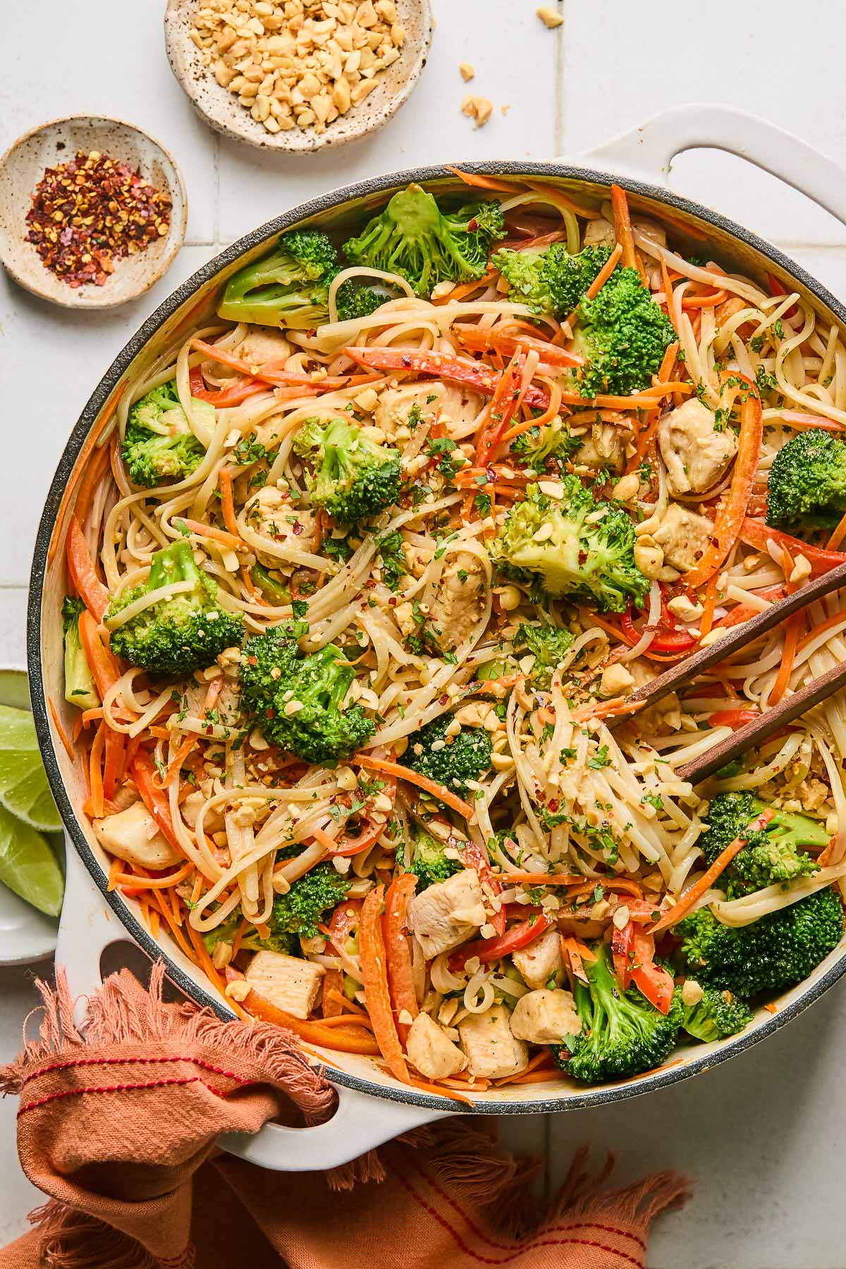 Peanut butter noodles with chicken and veggies in a large white skillet. It is garnished with cilantro and chopped peanuts and also in the pan is a pair of chopsticks. The handle of the pan is wrapped in a red towel and around the pan is a plate of lime wedges, a bowl of red pepper flakes, and a bowl of chopped peanuts.