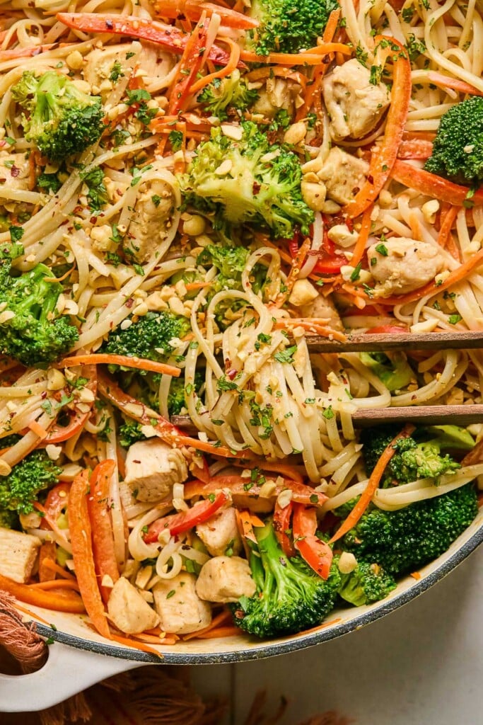 A pan of peanut butter Thai noodles with chicken and veggies. It is garnished with cilantro and chopped peanuts. A pair of chopsticks is wrapped around some of the noodles in the pan.