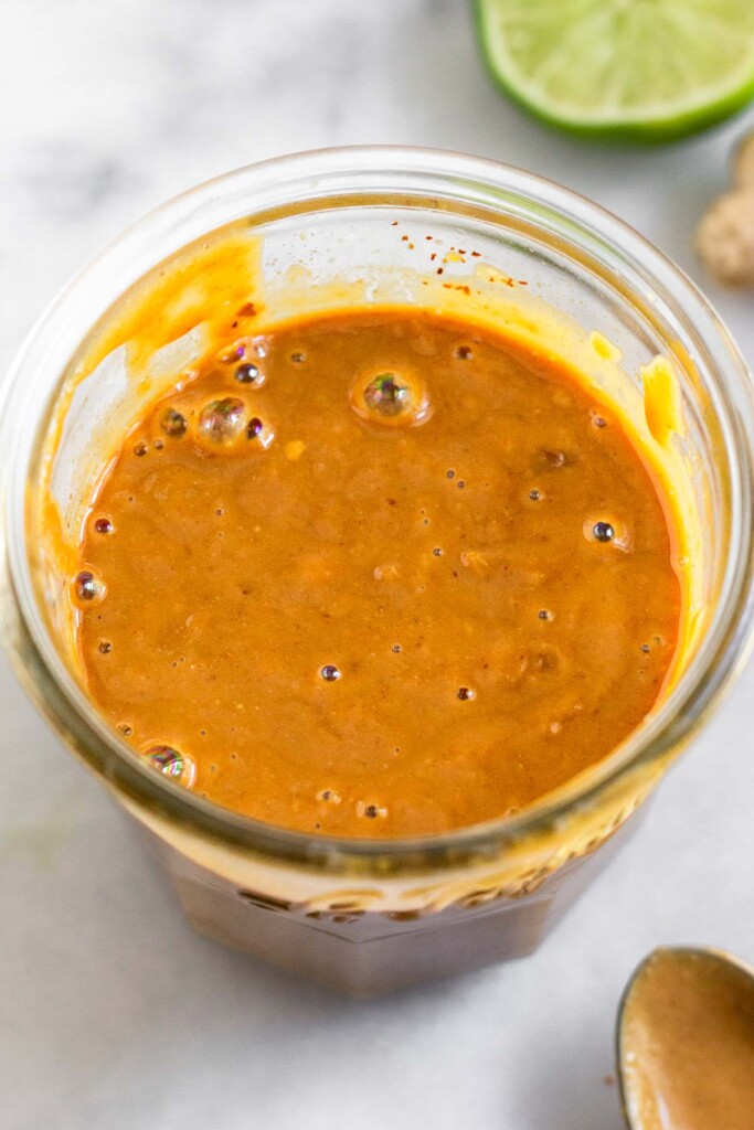 Peanut sauce in a medium jar. Around it is a spoon with more sauce on it, fresh ginger, and half a lime.