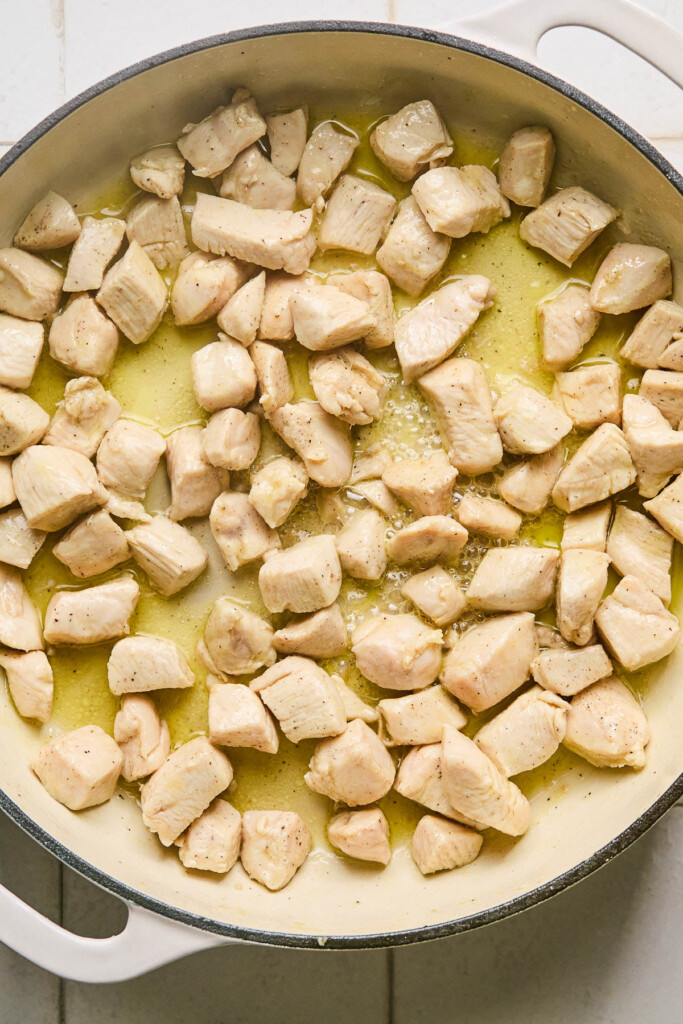 Large white skillet with bite sized pieces of cooked chicken in oil.