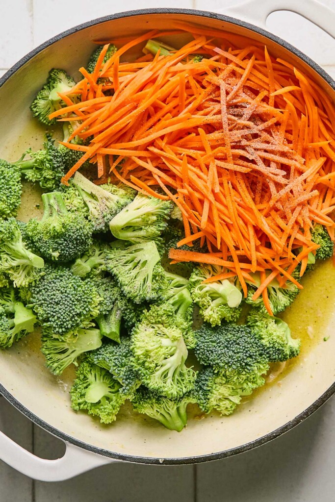 Large white skillet with broccoli florets and julienned carrots in oil before they are cooked.