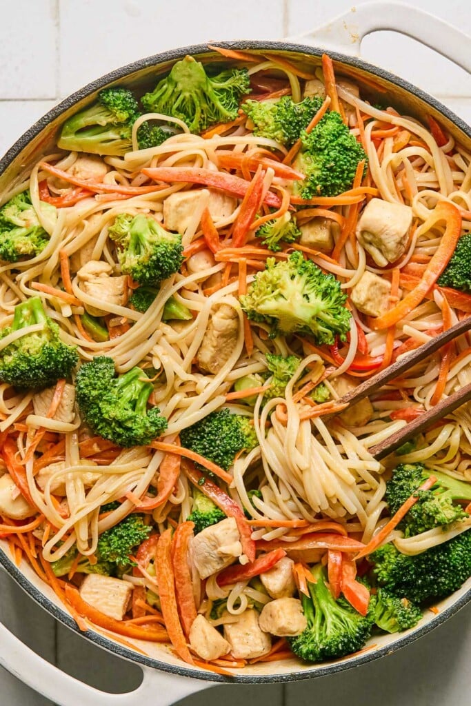 Large skillet with rice noodles, chicken, broccoli, carrots, and bell peppers. A pair of chopsticks is wrapped around some of the noodles in the pan.