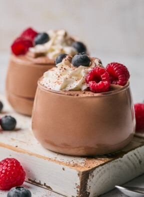 Two glass cups of protein pudding topped with whipped cream, fresh raspberries, fresh blueberries, and cinnamon. The cups are sitting on a white wooden board and there is more fresh fruit around them.