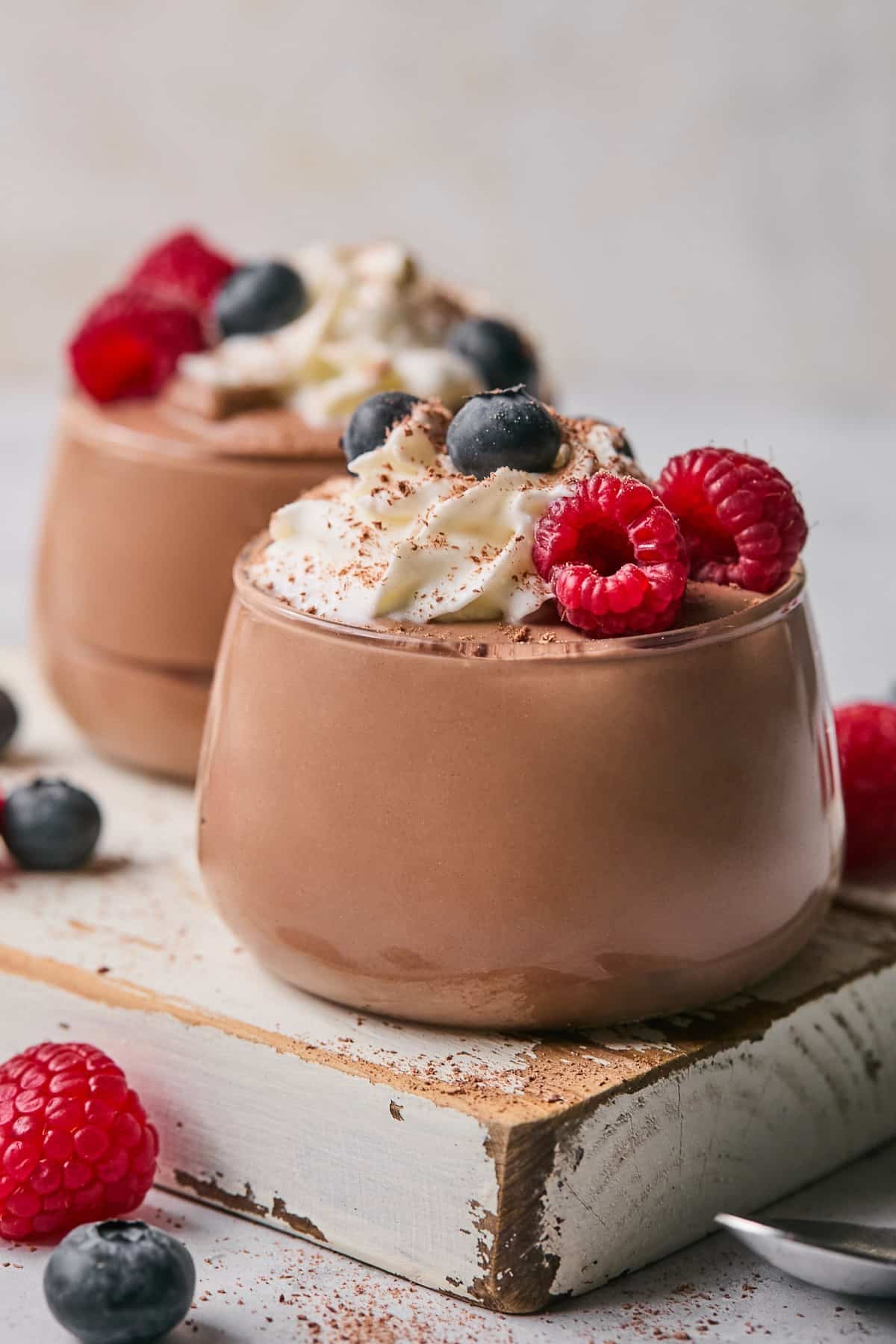 Two glass cups of protein pudding topped with whipped cream, fresh raspberries, fresh blueberries, and cinnamon. The cups are sitting on a white wooden board and there is more fresh fruit around them.