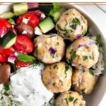 Greek chicken meatballs with herby rice, a greek salad with tomatoes, cucumbers, olives, and feta, and tzatziki sauce in a white bowl.