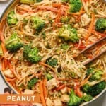 Peanut butter noodles with chicken and veggies in a large white skillet. It is garnished with cilantro and chopped peanuts and also in the pan is a pair of chopsticks. The handle of the pan is wrapped in a red towel and around the pan is a plate of lime wedges, a bowl of red pepper flakes, and a bowl of chopped peanuts.
