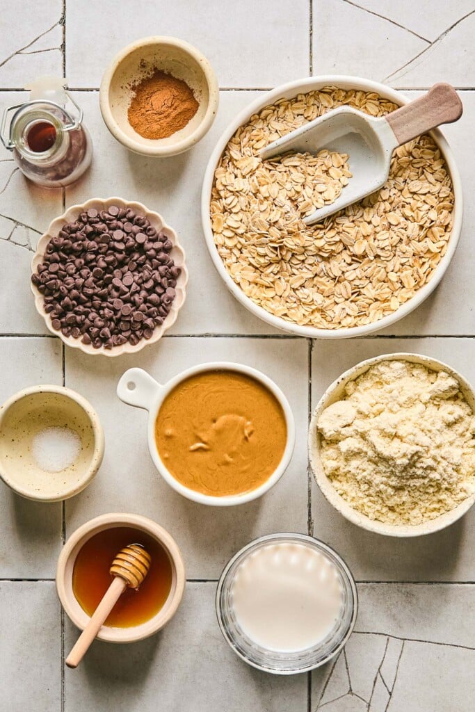 Grey tile counter with a bowl of rolled oats with a scoop in it, a bowl of vanilla protein powder, a measuring cup of peanut butter, a bowl of milk, a bowl of honey with a dipper in it, a small bowl of salt, a bowl of mini chocolate chips, a jar of vanilla extract, and a small bowl of cinnamon.