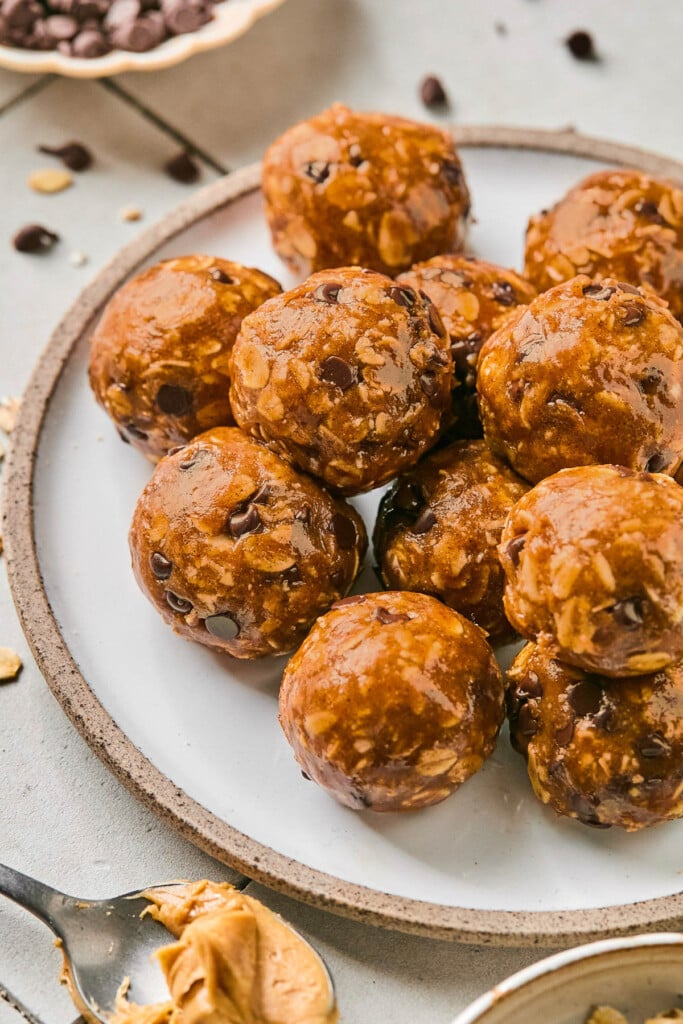 Peanut butter protein balls with oats and chocolate chips stacked on a white plate. Around the plate is a spoon full of peanut butter, scattered chocolate chips, and a bowl of chocolate chips.