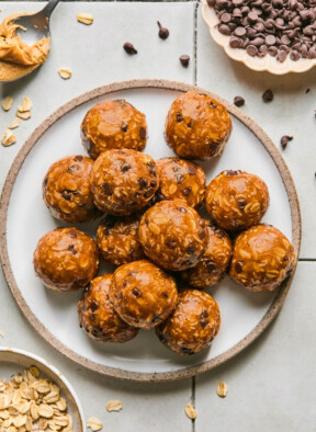 Peanut butter oatmeal protein balls stacked on top of each other on a plate. Around the plate is a bowl of rolled oats, a spoon with peanut butter on it, and a bowl of mini chocolate chips. Some oats and chocolate chips are on scattered around the plate.