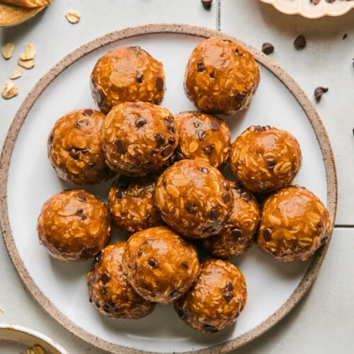 Peanut butter oatmeal protein balls stacked on top of each other on a plate. Around the plate is a bowl of rolled oats, a spoon with peanut butter on it, and a bowl of mini chocolate chips. Some oats and chocolate chips are on scattered around the plate.
