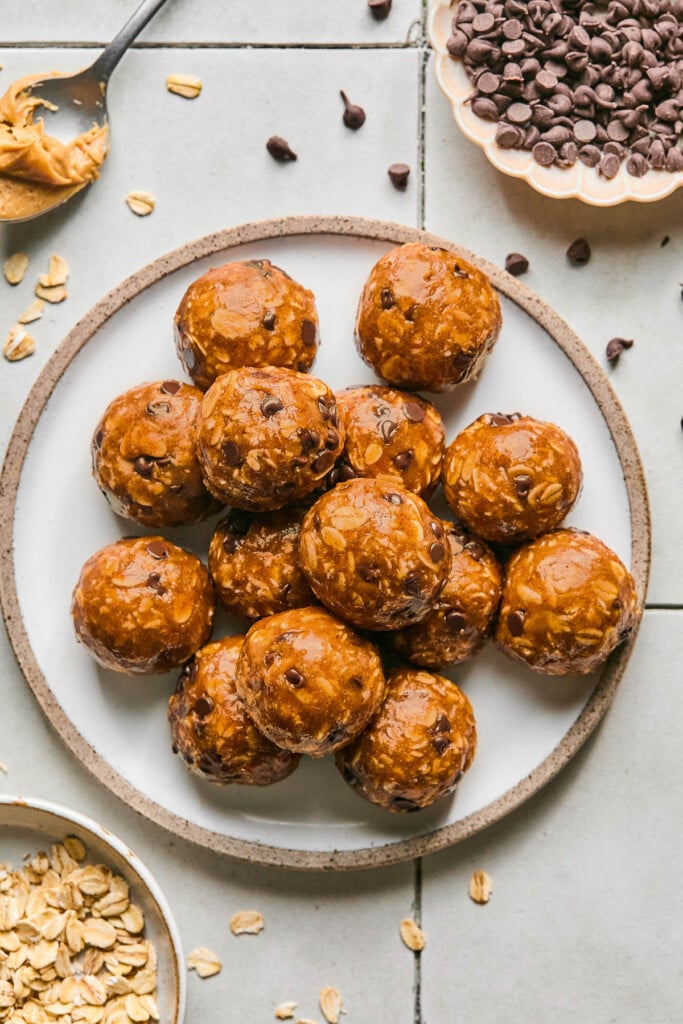 Peanut butter oatmeal protein balls stacked on top of each other on a plate. Around the plate is a bowl of rolled oats, a spoon with peanut butter on it, and a bowl of mini chocolate chips. Some oats and chocolate chips are on scattered around the plate.