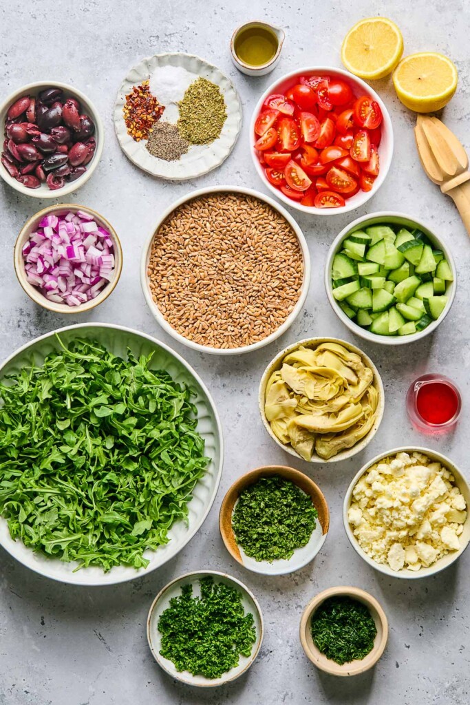 Grey concrete counter with a lemon cut in half with a wooden reamer next to it, a bowl of diced tomatoes, a bowl of diced cucumbers, a small dish of red wine vinegar, a bowl of artichoke hearts, a bowl of crumbled feta cheese, 3 small dishes of chopped herbs, a bowl of arugula, a bowl of uncooked farro, a bowl of diced red onions, a bowl of Kalamata olives, and plate of spices, and a small dish of olive oil.