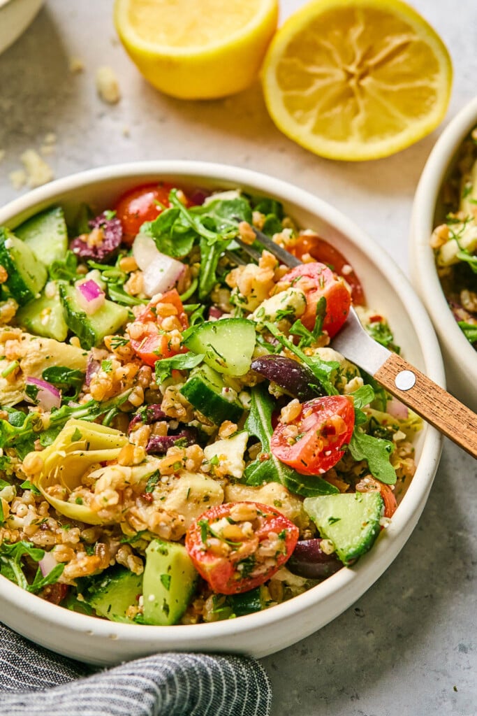 Greek farro salad in a bowl with a fork in it. Around the bowl is a lemon that has been cut in half, another bowl of farro salad, and a striped kitchen towel.