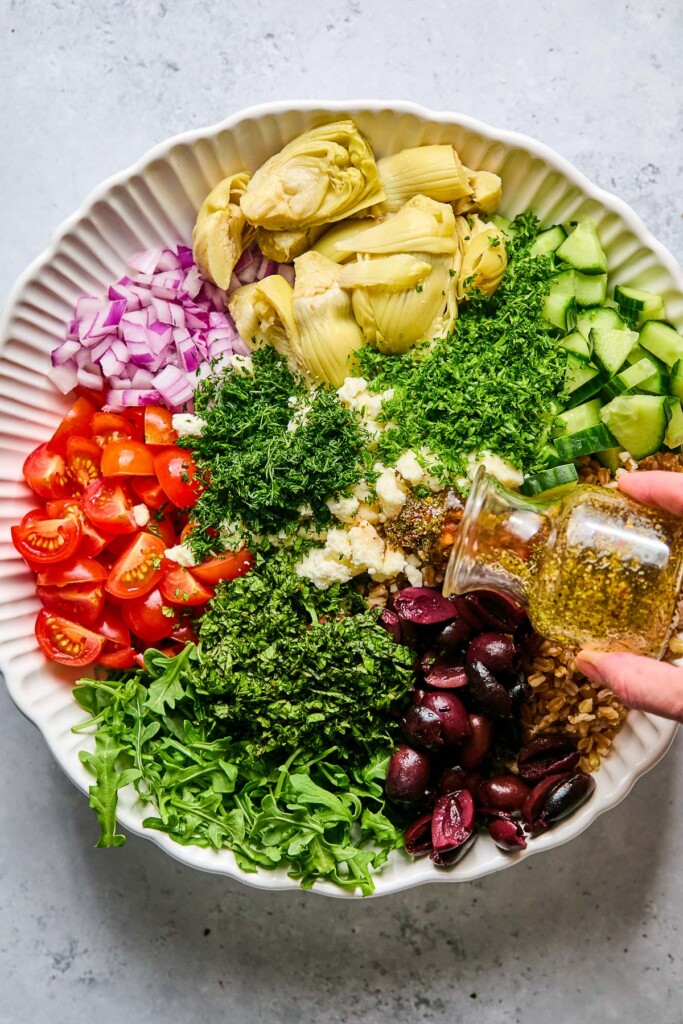 Large bowl with arugula, diced tomatoes, diced red onion, quartered artichoke hearts, diced cucumber, cooked farro, Kalamata olives, crumbled feta, and fresh herbs in it before it is mixed together. Dressing is being poured overtop of it.