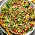 Large bowl filled with Mediterranean farro with two wooden salad tongs in the bowl. Around the bowl is a lemon cut in half, a striped linen towel, and a bowl with feta in it.
