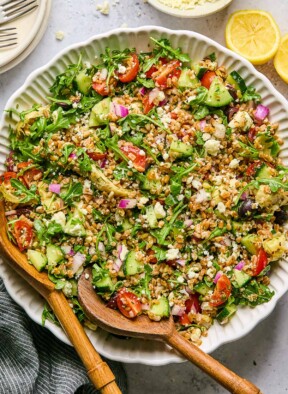 Large bowl filled with Mediterranean farro with two wooden salad tongs in the bowl. Around the bowl is a lemon cut in half, a striped linen towel, and a bowl with feta in it.