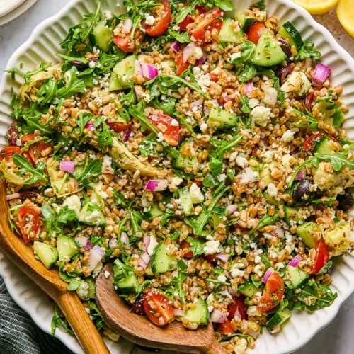 Large bowl filled with Mediterranean farro with two wooden salad tongs in the bowl. Around the bowl is a lemon cut in half, a striped linen towel, and a bowl with feta in it.
