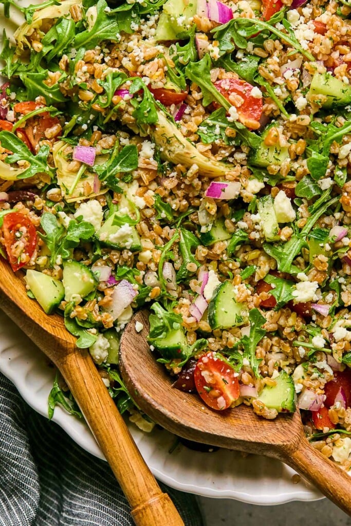 Farro salad with arugula, tomatoes, cucumbers, feta, olives, artichoke hearts, and herbs in a large bowl. Two wooden salad tongs are in the bowl as well.