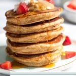 A stack of protein pancakes on a plate topped with yogurt, peanut butter, and strawberries with maple syrup dripping down them. Behind the plate is a bowl of diced strawberries and a jar of milk.