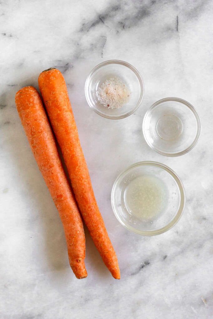 Marble counter with two medium carrots, a small bowl of salt, a small bowl of white vinegar, and a small bowl of coconut oil.