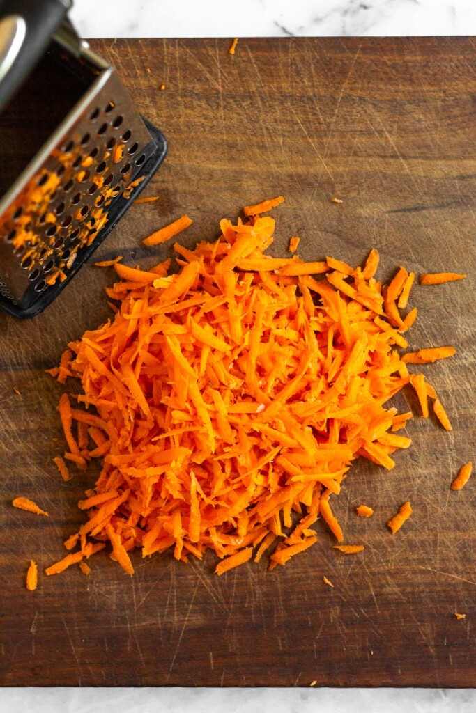 Wooden cutting board with a pile of shredded carrots and a box grater on it.