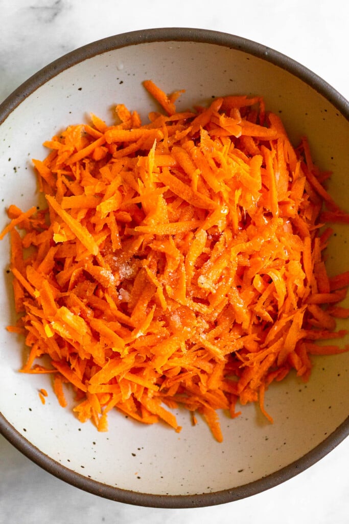 White bowl with shredded carrots with salt and oil sprinkled on top of them.
