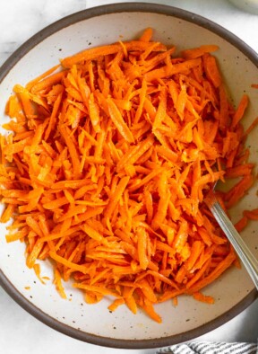 Raw carrot salad in a white bowl with a fork in it. Next to the bowl is a striped linen towel.