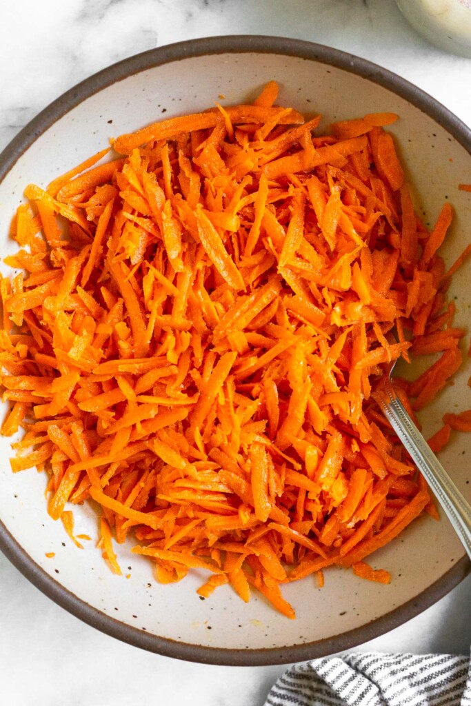 Raw carrot salad in a white bowl with a fork in it. Next to the bowl is a striped linen towel.