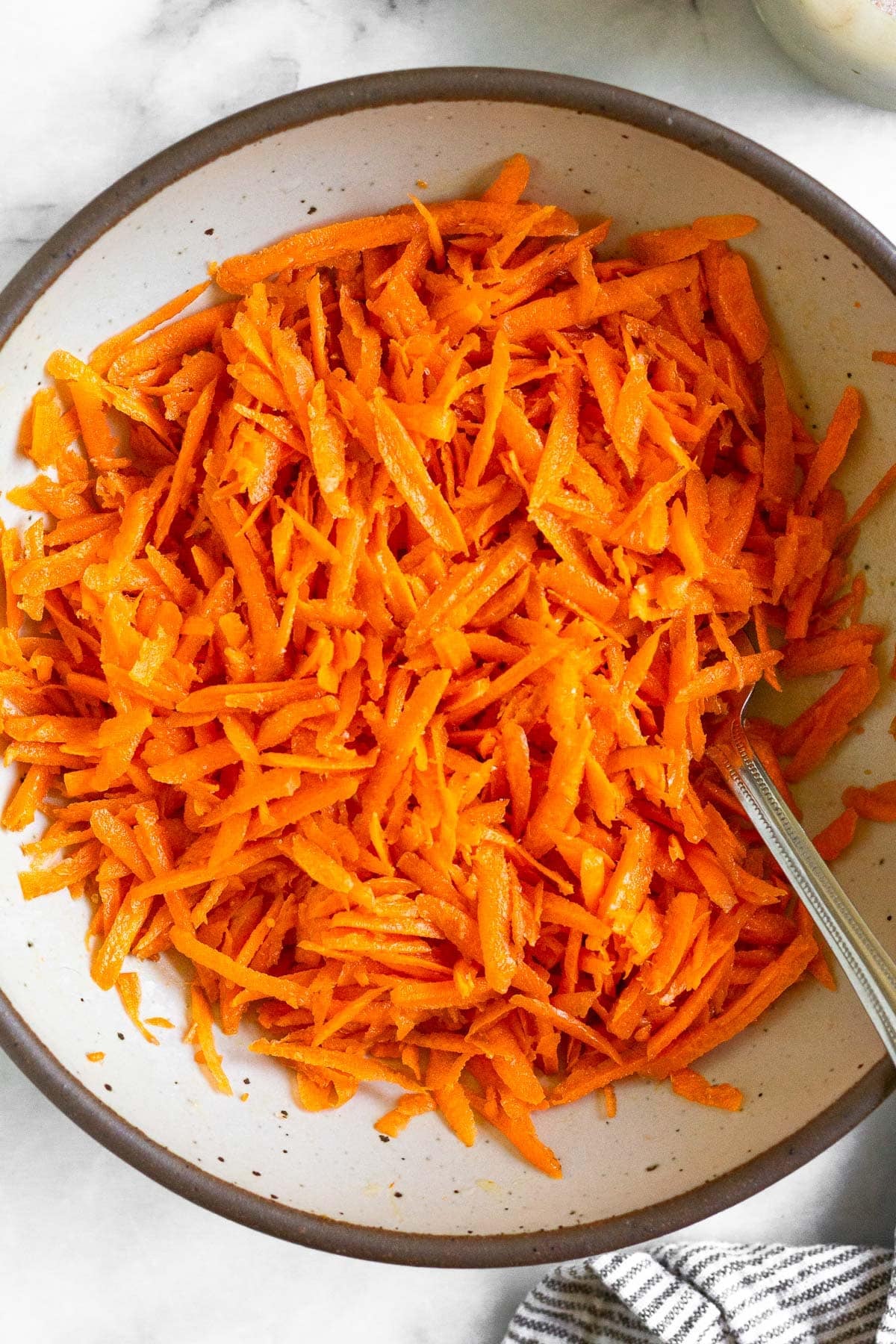 Raw carrot salad in a white bowl with a fork in it. Next to the bowl is a striped linen towel.