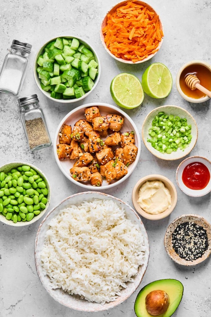 Grey concrete counter with a bowl of shredded carrots, a small bowl of honey with a honey dipper in it, a lime that has been cut in half, a bowl of diced green onions, a small bowl of sriracha, a small bowl of mayo, a bowl of black and white sesame seeds, half an avocado, a bowl of cooked white rice, a bowl of edamame, a bowl of cooked air fryer salmon bites, a salt and pepper shaker, and a bowl of diced cucumber.