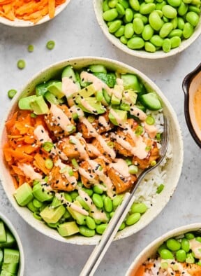 Salmon rice bowl with cubed salmon, white rice, edamame, diced avocado, shredded carrots, and diced cucumber with a fork in the bowl. Everything is drizzled with sriracha mayo and garnished with sesame seeds and green onions. Around the bowl is another rice bowl, a bowl of diced cucumber, a bowl of shredded carrots, a bowl of edamame, and a dish of sriracha mayo.