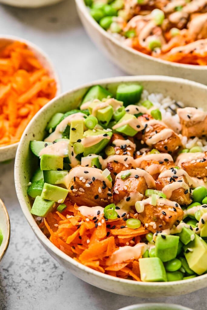 Air fryer salmon bowl with white rice, cubed salmon, edamame, diced avocado, shredded carrots, and diced cucumber. It is drizzled with sriracha mayo and garnished with sesame seeds and green onions. Behind the bowl is a bowl of shredded carrots and another salmon bowl.