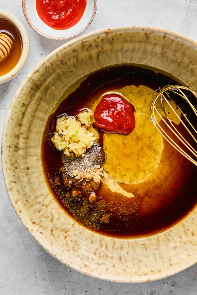 Bowl filled coconut aminos, honey, sriracha, minced garlic, and spices with a whisk in it. Next to it is a small bowl of sriracha and a small bowl of honey.