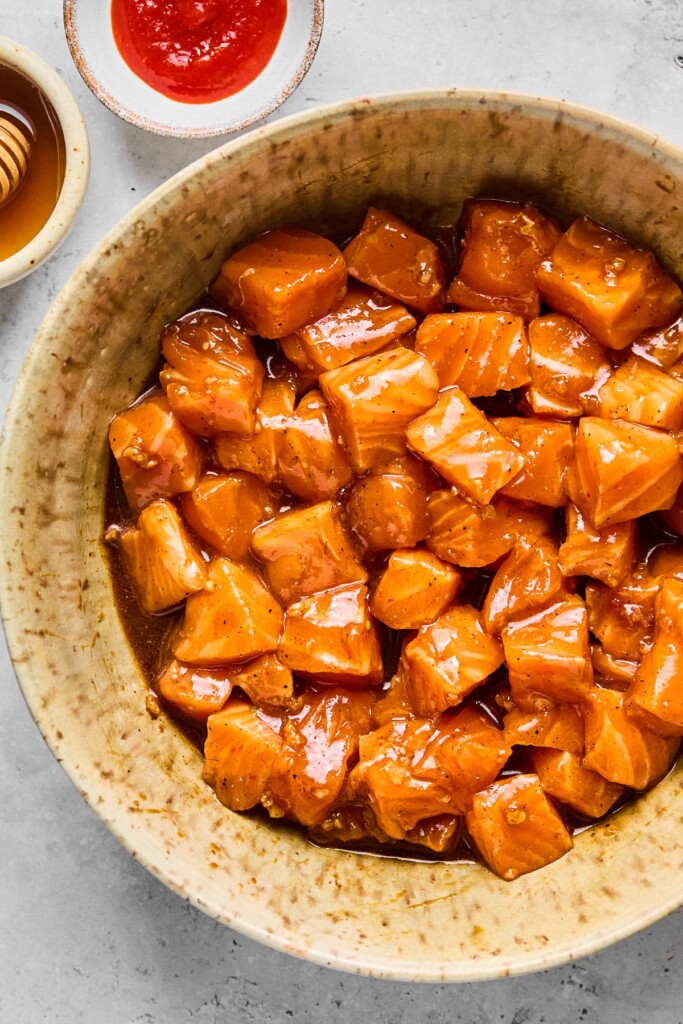 Cubed raw salmon coated in a brown marinade in a bowl. Next to the bowl is a small bowl of sriracha and a small bowl of honey.