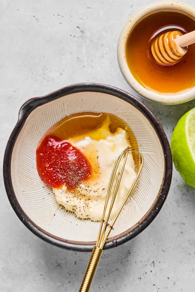 Small bowl of mayo, sriracha, honey, lime juice, salt, and pepper before it is mixed together. There is a small whisk in the bowl. Around the bowl is half a lime and a small dish of honey with a honey dipper in it.