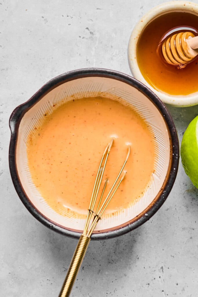 Small bowl of sriracha mayo with a small whisk in it. Around the bowl is half a lime and a small dish of honey with a honey dipper in it.