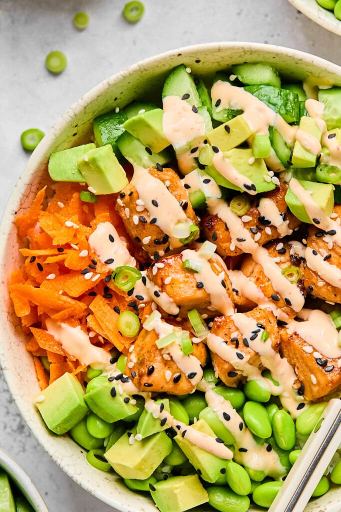 Close up of a salmon and rice bowl with edamame, diced avocado, shredded carrots, and diced cucumber. It is topped with sriracha mayo, sliced green onions, and sesame seeds.