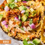 Bbq chicken tacos in a corn tortilla with coleslaw, shredded bbq chicken, avocado, shredded cheese, red onion, and cilantro on a plate.