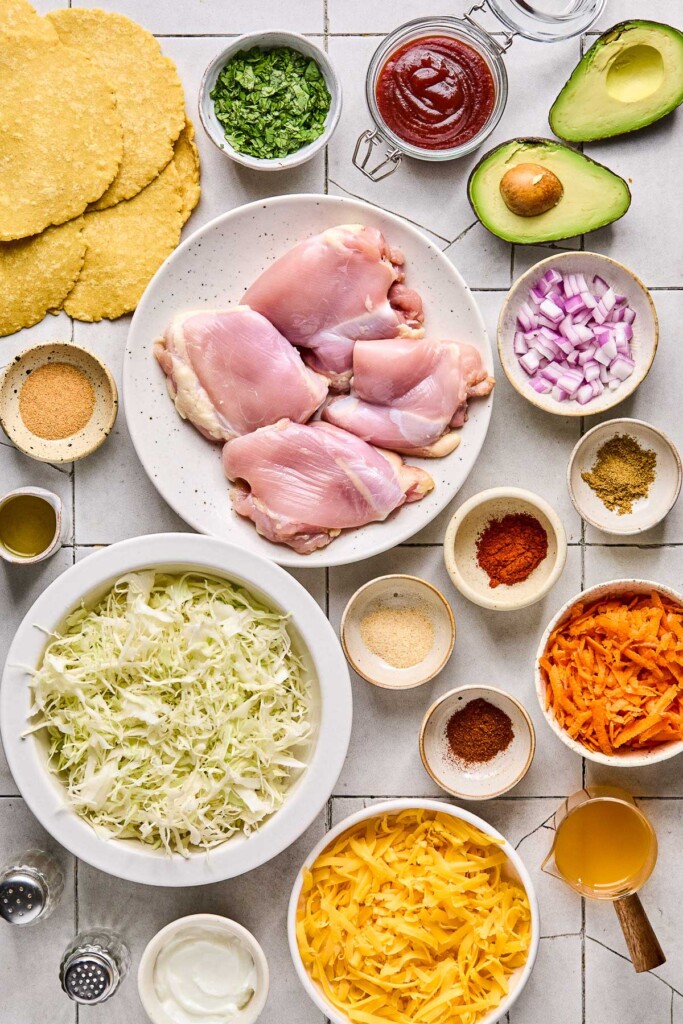 Grey tile counter with a jar of bbq sauce, an avocado that has been cut in half, a bowl of diced red onion, four small bowls with different spices in them, a bowl of shredded carrots, a measuring cup of apple cider vinegar, a bowl of shredded cheddar cheese, a small bowl of greek yogurt, salt and pepper shakers, a bowl of shredded cabbage, a small jar of olive oil, a small bowl of garlic powder, a pile of corn tortillas, and a bowl of cilantro.