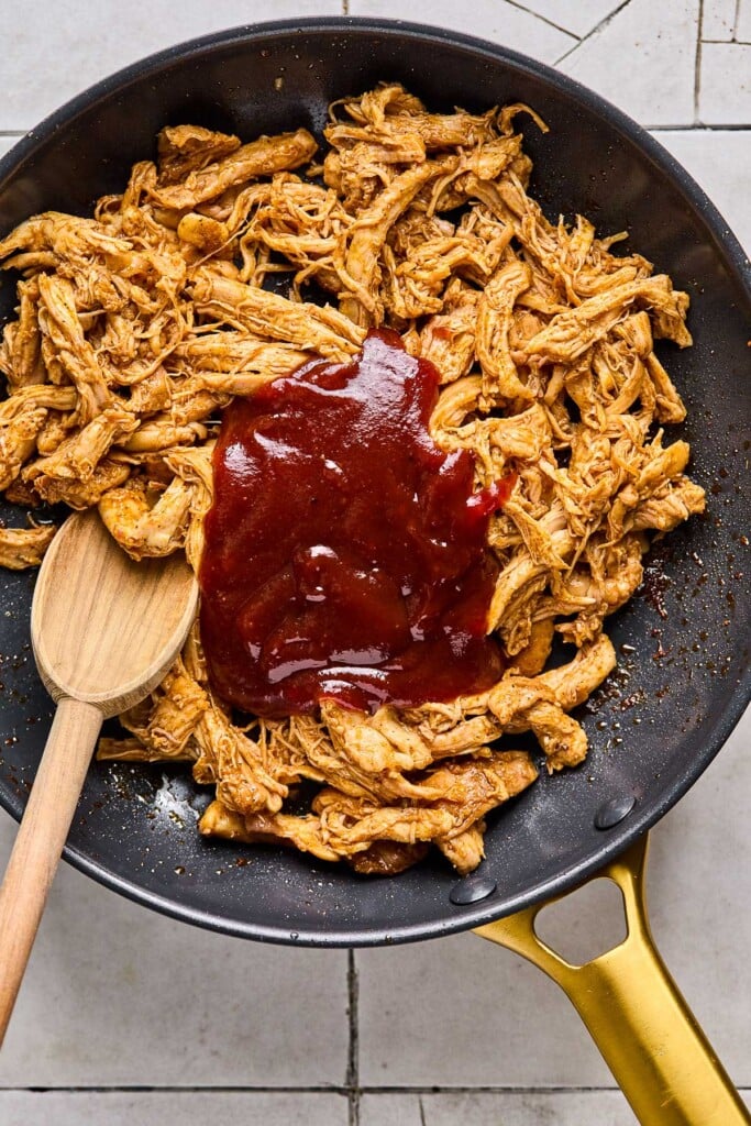 Shredded chicken in a pan with a pile of barbeque on it before it is mixed together. A wooden spoon is also in the pan.
